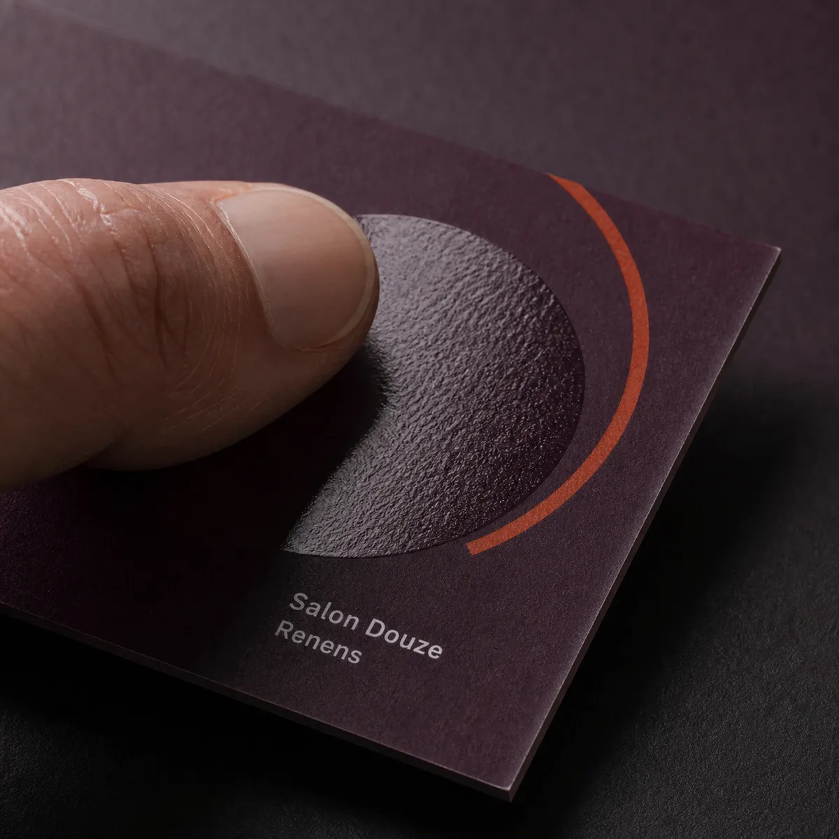 Pouce glissant sur une carte prune mate et s'arrêtant sur un disque verni brillant, éclairé par une lumière rasante - carte de visite vernis sélectif